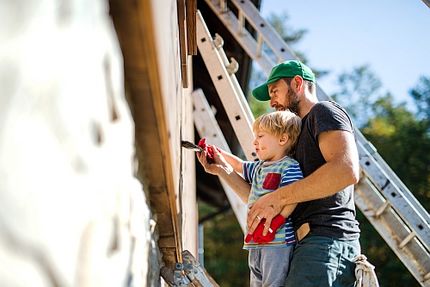 Vater streicht mit Sohn im Kindergartenalter die Fassade eines alten Hauses.