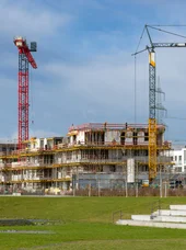 Lehrgang Städtebaurecht Im Vordergrund befindet sich eine große, freie Wiesenfläche. Im Hintergrund sind Gebäude und Kräne zu erkennen.