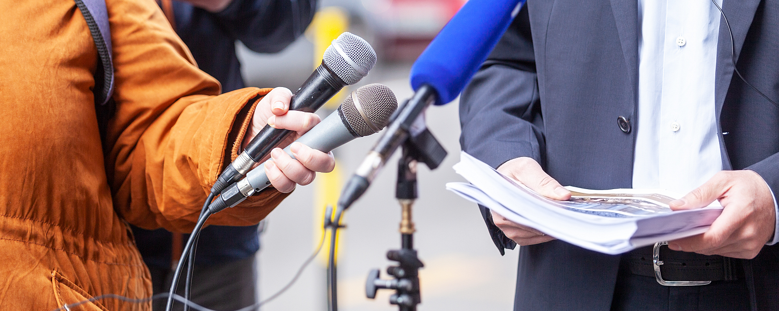 Foto: Interviewsituation, im Vordergrund Mikrofone, dahinter Interviewter im Anzug mit Dokumenten in den Händen.© wellphoto/stock.adobe.com